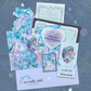 A flat lay of pastel blue and lavender craft paper embellishments, stickers, and a small printed card arranged on a textured blue-gray background for a snail mail club. The designs feature whimsical fairy and floral hydrangea illustrations, with text including 'i dreamt in full bloom' and 'sea milk.'