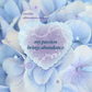 A monthly affirmation card featuring a central heart-shaped design with the text 'my passion brings abundance' in dark teal, surrounded by white decorative swirls. The background is a soft-focus photograph of light blue and lavender hydrangea flowers.