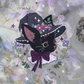 A decorative sticker design featuring a black cat wearing a witch hat adorned with pink and purple flowers, green leaves, and white stars, with a dark purple bow. The sticker is shown on a soft lavender floral background.