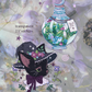 A styled flat-lay of two decorative stickers on a soft lavender floral background. One sticker shows a black cat wearing a witch hat with flowers and a purple bow; the other shows a round illustrated potion bottle with a moon lid and botanical terrarium inside.