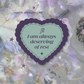 A monthly affirmation card featuring a central scalloped heart shape with the text 'I am always deserving of rest' in dark purple lettering on a pale mint background. The card is set against a soft-focus background of lavender and purple flowers, with small decorative stars and a spiral motif.